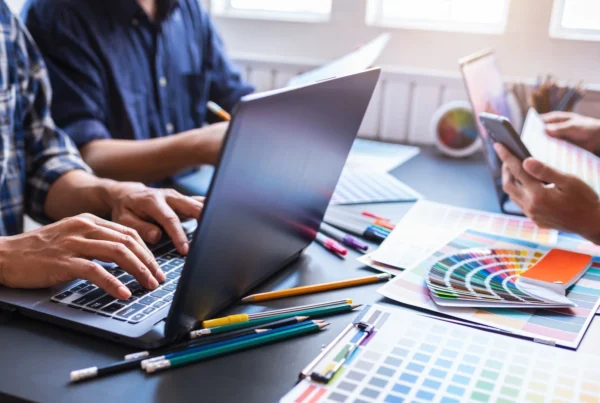 Designers working at a desk with laptops, colour swatches, pencils, and printed colour palettes while discussing colour choices.