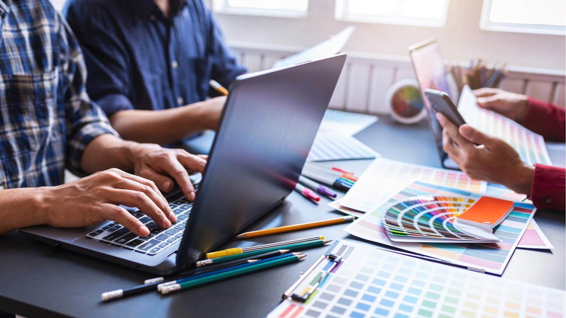 Designers working at a desk with laptops, colour swatches, pencils, and printed colour palettes while discussing colour choices.